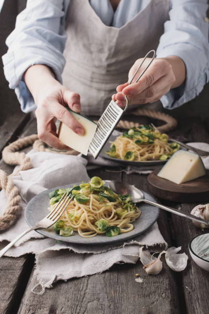 Woman cooking soy noodles, fried garlic, Brussels sprouts and chesse in a serving plate on a cutting board, forks and spoons vegan concept in a rustic style stock photo