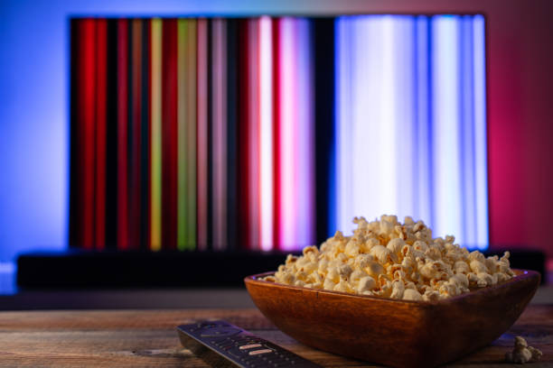 A wooden bowl of popcorn and remote control in the background the TV works. Evening cozy watching a movie or TV series at home A wooden bowl of popcorn and remote control in the background the TV works. Evening cozy watching a movie or TV series at home. popcorn snack bowl isolated stock pictures, royalty-free photos & images