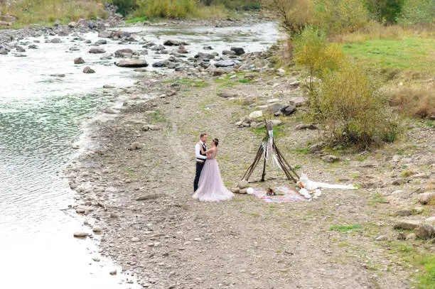 An attractive newlywed couple, a happy and joyful moment. Man and woman hug and kiss in holiday clothes. Wedding Cermonia with Boho-style décor by the river in the fresh air.
