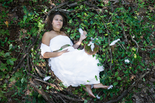 Fantasy portrait of a pregnant Caucasian woman wearing a white gown lying curled up in a giant nest of grapevine, branches and leaves outdoors nesting while she waits for her baby to be born