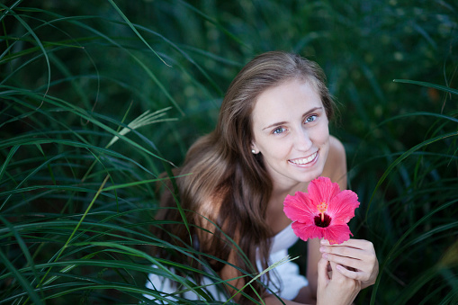 Portrait of a smiling girl with long brown hair holding a pink hibiscus flower in the middle of tall pampas grass outdoors