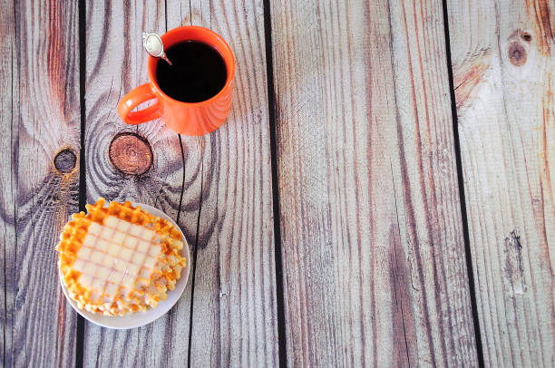 An orange ceramic mug of hot coffee and a plate with Parisian waffles and creamy syrup.