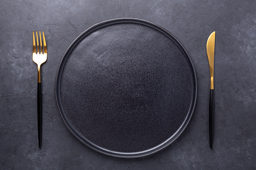 Dark table setting. Empty black ceramic plate, knife and fork on stone background - Image