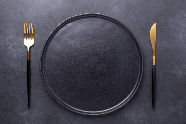 Dark table setting. Empty black ceramic plate, knife and fork on stone background stock photo