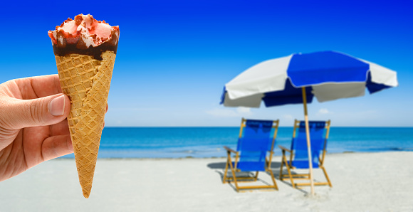 hand holding a fresh strawberry flavor ice cream cone in front of blurred sun loungers and a beach umbrella on silver sand