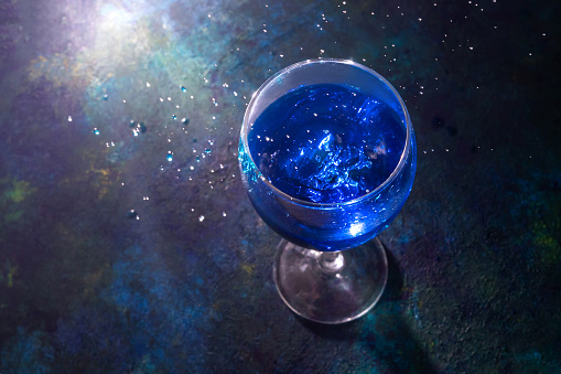 Blue cocktail in wineglass with splash on dark background with back light.