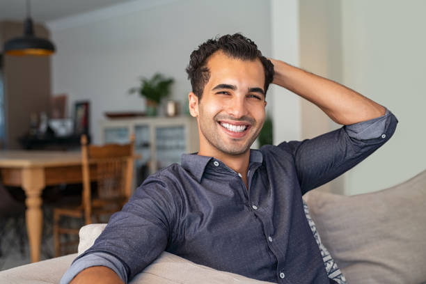 Handsome indian guy smiling Happy successful young businessman relaxing on a comfortable sofa. Portrait of smiling indian man looking at camera sitting on couch while run his hand through his hair. Stylish middle eastern guy relaxing at home. charming photos stock pictures, royalty-free photos & images