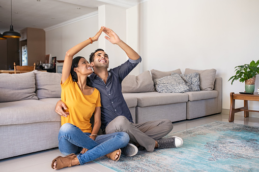 Handsome young man with beautiful indian woman dreaming a new home. Happy young married couple moves to new apartment with copy space. Happy middle eastern couple making roof with hands symbol of new home and insurance protection plan.
