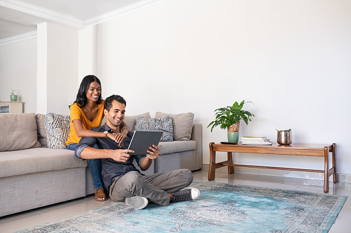 Young middle eastern couple using digital tablet at home while sitting on couch and floor. Indian woman embracing from behind her boyfriend while watching video on digital tablet at home. Happy smiling couple in video call conference, copy space.