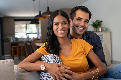 Portrait of happy young indian couple relaxing together on couch. Middle eastern couple cuddling on couch at home in the living room while looking at camera. Portrait of lovely man embracing from behind his beautiful girlfriend sitting in sofa.