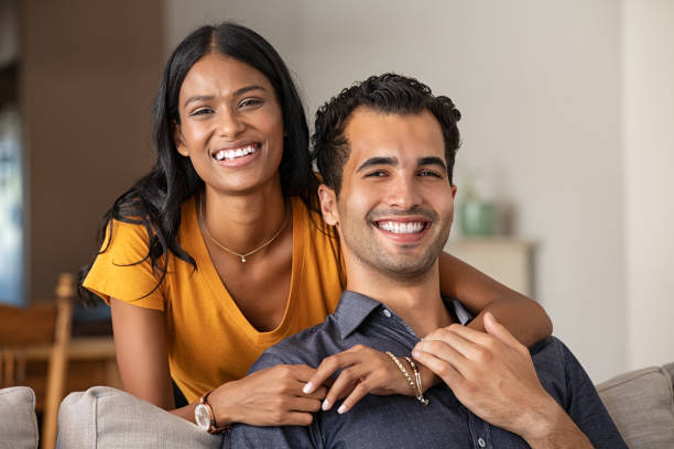 Happy indian couple at home portrait Smiling indian woman hugging her husband on the couch from behind at home. Loving middle eastern couple looking at camera with big grin in the living room. Portrait of laughing girl embracing handsome latin man on sofa. south asia stock pictures, royalty-free photos & images