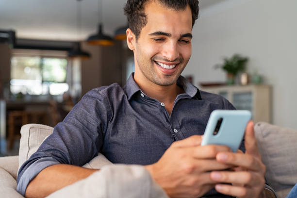 Latin man using smartphone at home Cheerful businessman using smartphone while sitting on sofa at home. Handsome young indian man sitting on couch reading messages on mobile phone. Hispanic guy working from home with cellphone. person on phone stock pictures, royalty-free photos & images