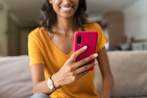 Close up hands of young middle eastern woman using cellphone at home. Smiling latin girl in video call with her smartphone indoor. Young indian woman sitting on sofa using app and messaging with mobile phone with pink cover.