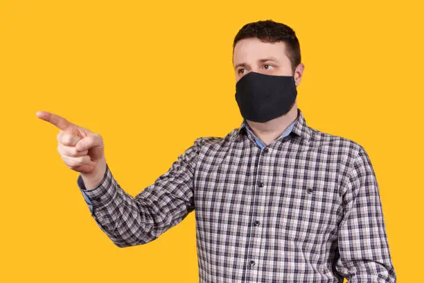 Photo of Man in black face mask pointing with finger, isolated on orange background.