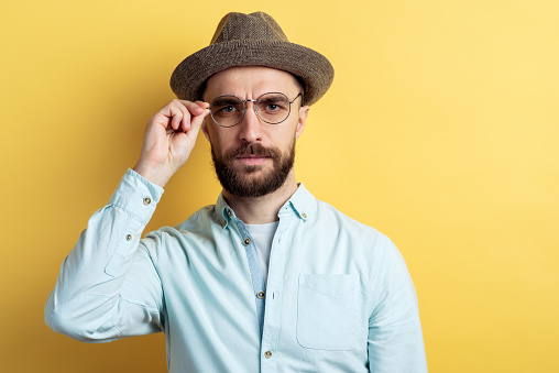 Portrait of brunette man with beard wearing in shirt, hat and glasses. He frown eyesbrows and looking at the camera touching glasses
