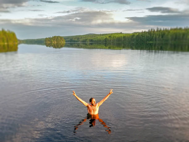 homem saudável está tomando banho frio no lago e está animado, mãos para cima e olhando para o pôr do sol no norte da suécia - sweden summer swimming lake - fotografias e filmes do acervo