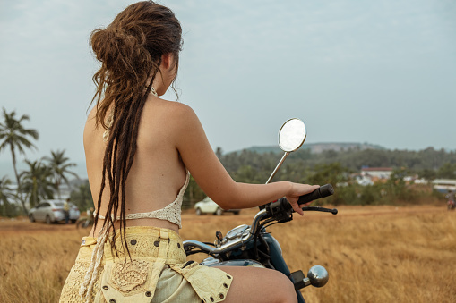 A woman sits on a motorcycle in a field waiting for sunset, back view