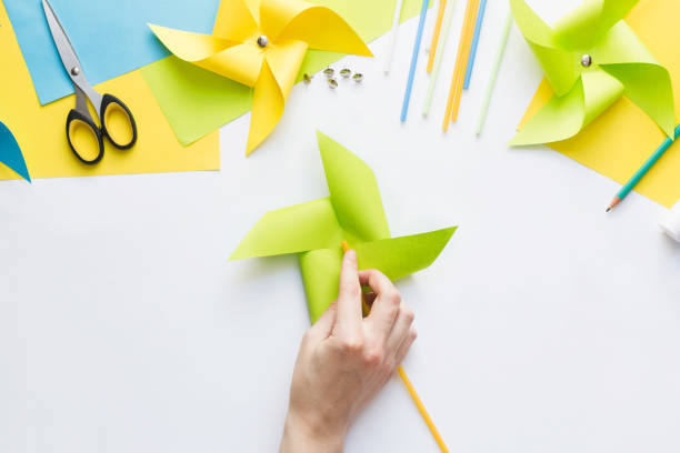 istock How to make paper green windmill toy with children at home. Step by step instructions. Hands making DIY summer project. Top view. Step 11. Turn windmill to other side and attach straw to button. 1223364375
