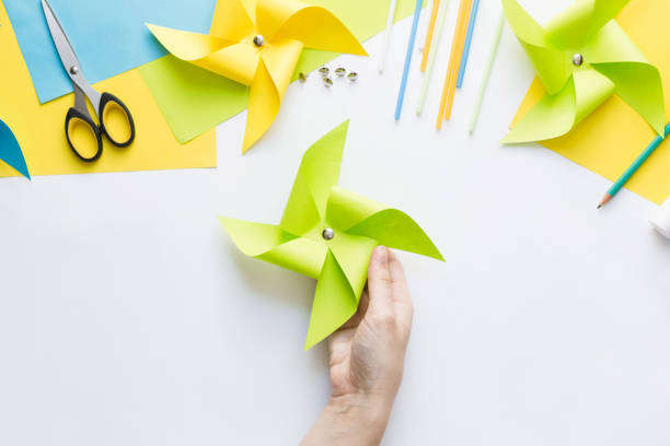 How to make paper green windmill toy with children at home. Step by step instructions. Hands making DIY summer project. Top view. Step 10. Attach office button to mark center.