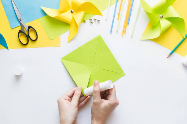 istock How to make paper green windmill toy with children at home. Step by step instructions. Hands making DIY summer project. Top view. Step 6. Apply glue to right corner of resulting triangle cut out. 1223364460