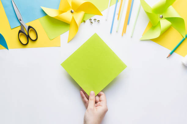 istock How to make paper green windmill toy with children at home. Step by step instructions. Hands making DIY summer project. Top view. Step 1. Take a square sheet of colored paper. 1223364550