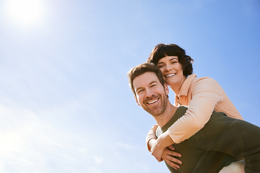 Portrait of a smiling young man carrying his wife on his back against a blue sky while having fun outside together on a sunny day