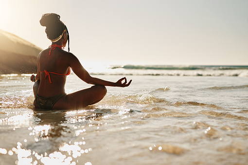 Full length shot of an unrecognizable woman sitting alone on the beach and meditating