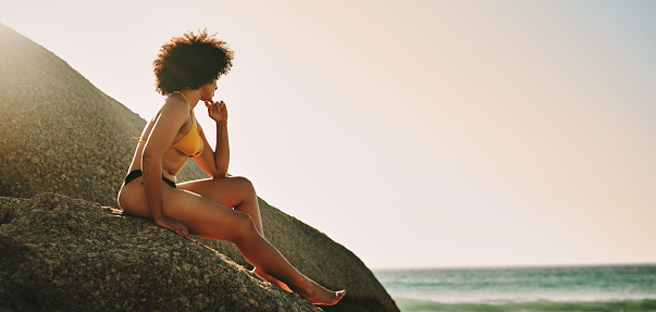 Full length shot of an unrecognizable woman sitting on a rock and posing during a day out at the beach