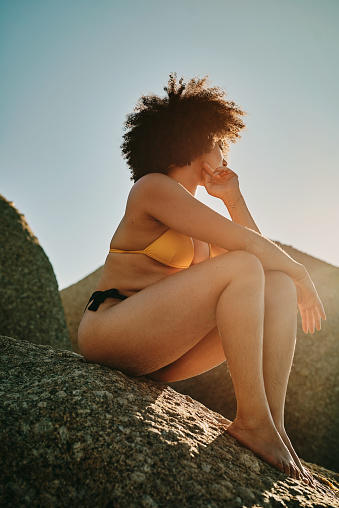 Full length shot of an unrecognizable woman sitting on a rock and posing during a day out at the beach