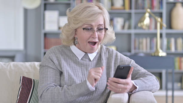 istock Old Woman Celebrating Success on Smartphone on Sofa 1223373670
