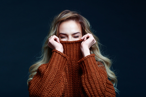 Portrait of long hair blond young woman wearing brown sweater, covering her face. Studio shot against black background.