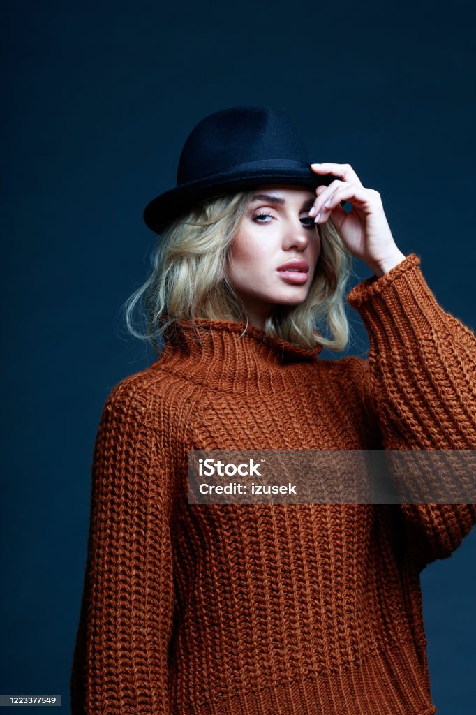 Autumn portrait of elegant woman in brown sweater Fashion portrait of long hair blond young woman wearing brown sweater and black hat, looking at camera. Studio shot against black background. Sale Stock Photo