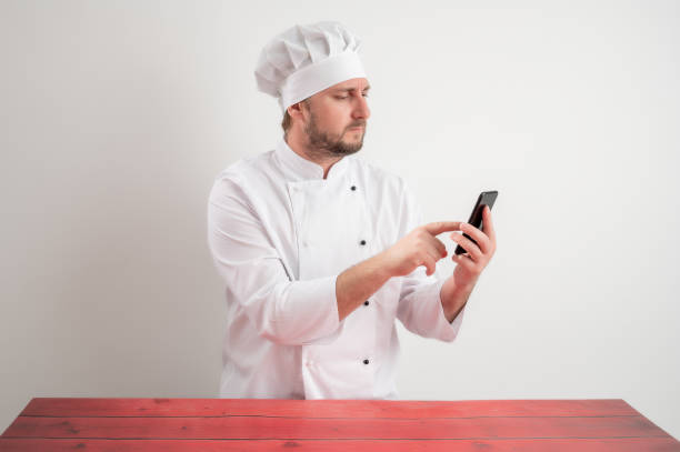 istock Young male chef in white uniform holding smartphone, text messages 1223379606