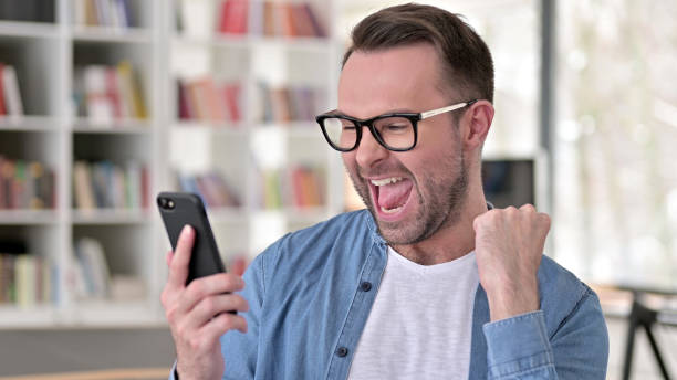 istock Young Man in Glasses Celebrating Success on Smartphone 1223380656