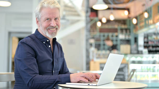 istock Old Man with Laptop Smiling at Camera in Cafe 1223381433