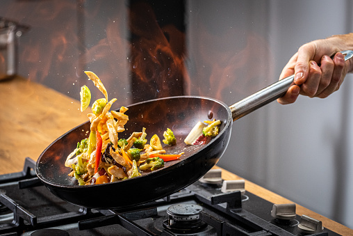 Chef tossing flaming vegetable in a frying pan.
