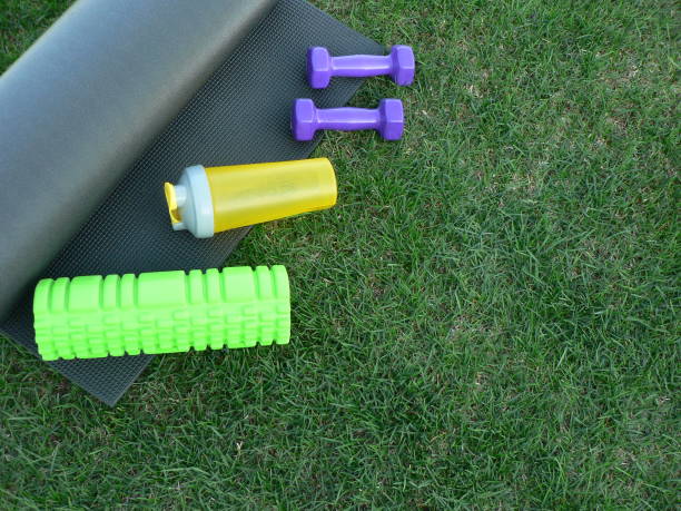 Keeping fit and exercising outdoor or at home. Purple dumbbells, yellow bottle for water, green foam roll on a yoga mat on green grass lawn in a backyard or park. Healthy lifestyle. Copy space. mantenerse en forma y hacer ejercicio al aire libre o en casa. mancuernas púrpuras, botella amarilla para el agua, rollo de espuma verde en una estera de yoga en césped de césped verde en un patio trasero o parque. estilo de vida saludable. copiar espac