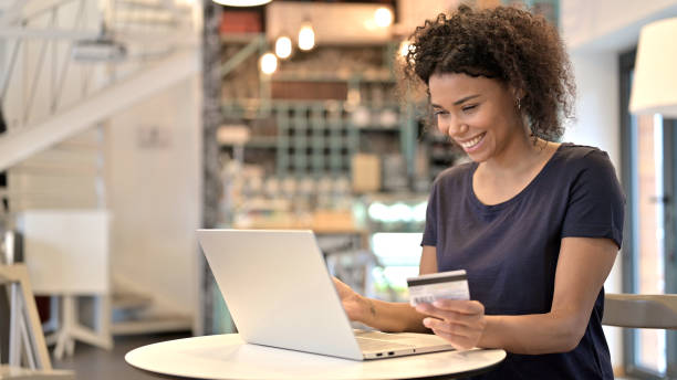 успешная онлайн-оплата на ноутбуке молодой африканской женщиной, кафе - credit card women laptop electronic banking стоковые фото и изображения