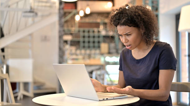 Young African Woman Reacting to Loss on Laptop in Cafe Young African Woman Reacting to Loss on Laptop in Cafe shocked computer stock pictures, royalty-free photos & images