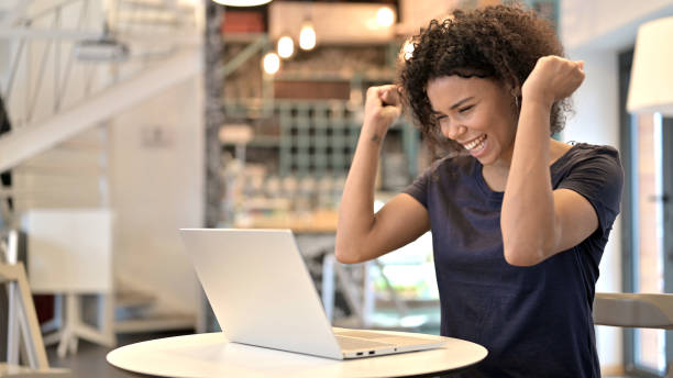 joven mujer africana celebrando el éxito en el ordenador portátil en el café - searching for success fotografías e imágenes de stock
