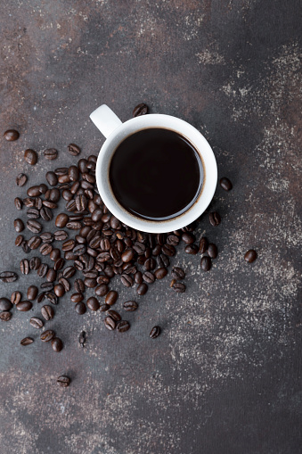 Direct above view of roasted coffee beans and white coffee cup with black coffee on dark brown background.