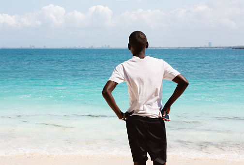 Young african man, 23 years old, standing at the beach of an island, holding his mobile phone in his hand,  in the background you can see the skyline of Dar es Salaam, Tanazania