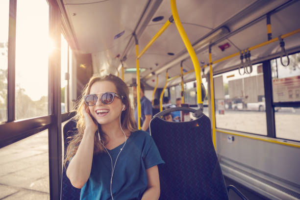 Smiling mid adult woman wearing earphone talking on mobile phone in bus. Happy female is commuting by public transport on sunny day.