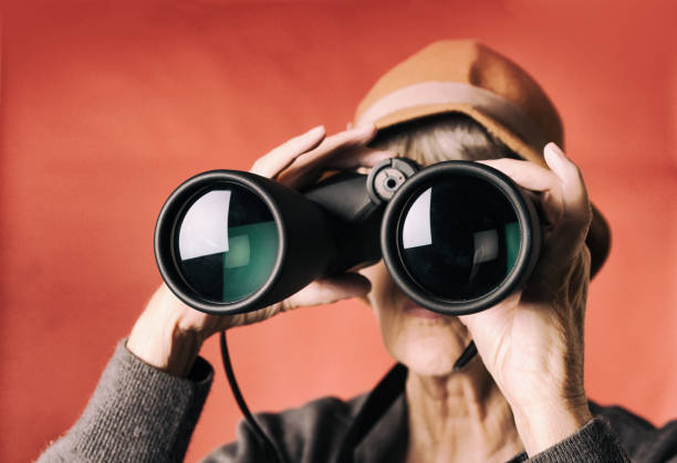 Senior woman peers curiously through binoculars, A senior woman peeps through large binoclurs which obscure her face. coral colored photos stock pictures, royalty-free photos & images
