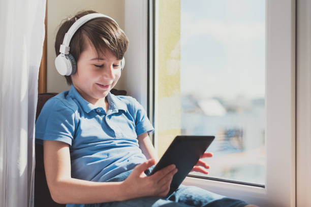 istock Boy wearing headphones using tablet sitting on pillow and windowsill. Soft focus. Quarantine and online school concept 1223399602