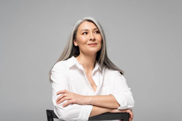 bella donna asiatica sognante con i capelli grigi seduti su sedia isolata sul grigio - capelli lunghi foto e immagini stock