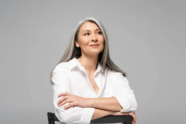 beautiful dreamy asian woman with grey hair sitting on chair isolated on grey