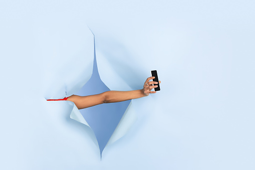 Female hand giving smartphone in torn blue paper hole background. Breaking on, breakthrought with gesturing. Concept of celebration, shopping, proposal, sales, ad. Copyspace. Promotions, discounts.