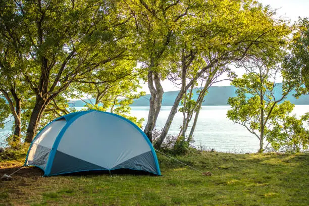 View Of Blue Small Tent Pitched On Shaded By Trees Area Near Lake And Mountains.