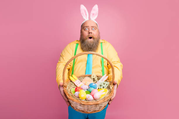 ritratto del suo bel stupito impressionato funky felice ragazzo barbuto tenendosi in mano dandoti uno spuntino da panificio da basket artigianale pasquale festale isolato su sfondo color pastello rosa - costume da coniglietto foto e immagini stock